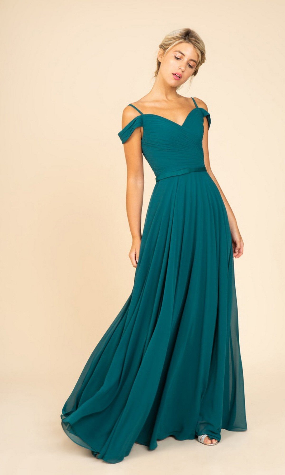 A woman stands against a beige background wearing the Long Prom Dress: Eureka 7611—an off-the-shoulder chiffon gown with a ruched fitted bodice, lace-up open back, and flowing teal floor-length skirt, paired with silver open-toed shoes.